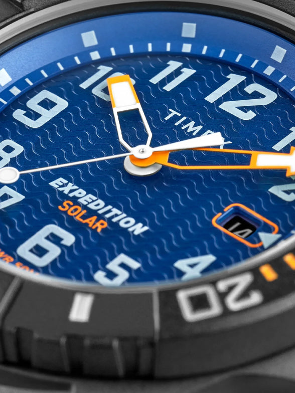 A macro shot of a Timex Expedition Solar watch with a deep blue textured dial, featuring white and orange accents. The dial includes bold numerals, a date window, and a rugged black bezel with markings, emphasizing its durable and sporty design.