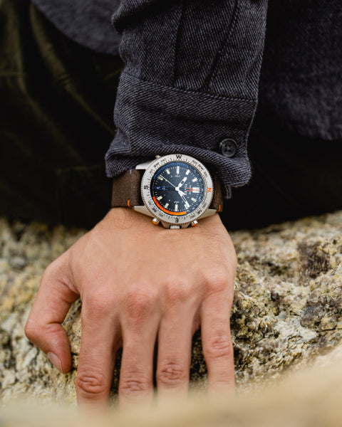 A person wearing a Timex expedition-style wristwatch with a black dial and a brown leather strap while resting their hand on a rocky surface. The watch has a sturdy build, compass markings on the bezel, and is designed for outdoor exploration.