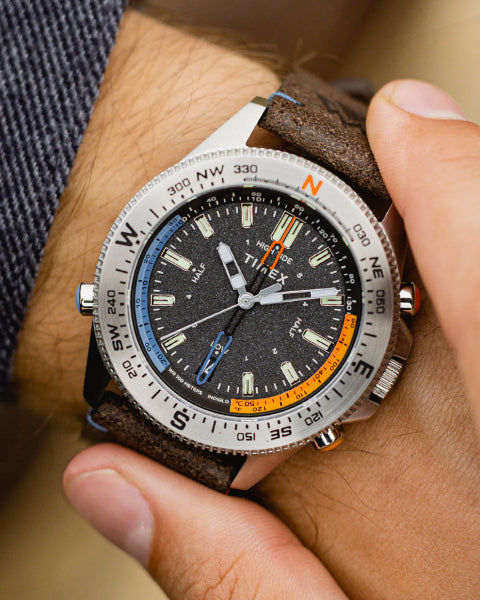 A close-up of a person adjusting their Timex expedition wristwatch. The watch has a rugged black dial with tide and compass indicators, luminescent hands, and a textured brown leather strap, reflecting an adventurous and exploration-ready design.
