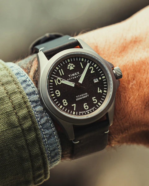 close-up of a Timex Titanium Automatic wristwatch on a person's wrist. The watch features a black dial with white numerals, a date display, and a durable titanium case. It is paired with a black leather strap, complementing the rugged and adventure-ready aesthetic. The wearer is dressed in a green and denim jacket, adding to the outdoorsy style.