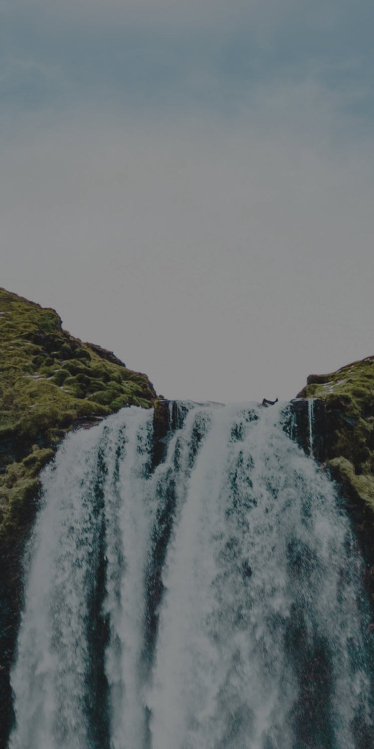 A scenic waterfall cascading down a moss-covered cliffside, surrounded by lush green hills. The water flows powerfully over the edge, creating a misty effect. The background features a bright sky, adding to the serene and natural beauty of the landscape